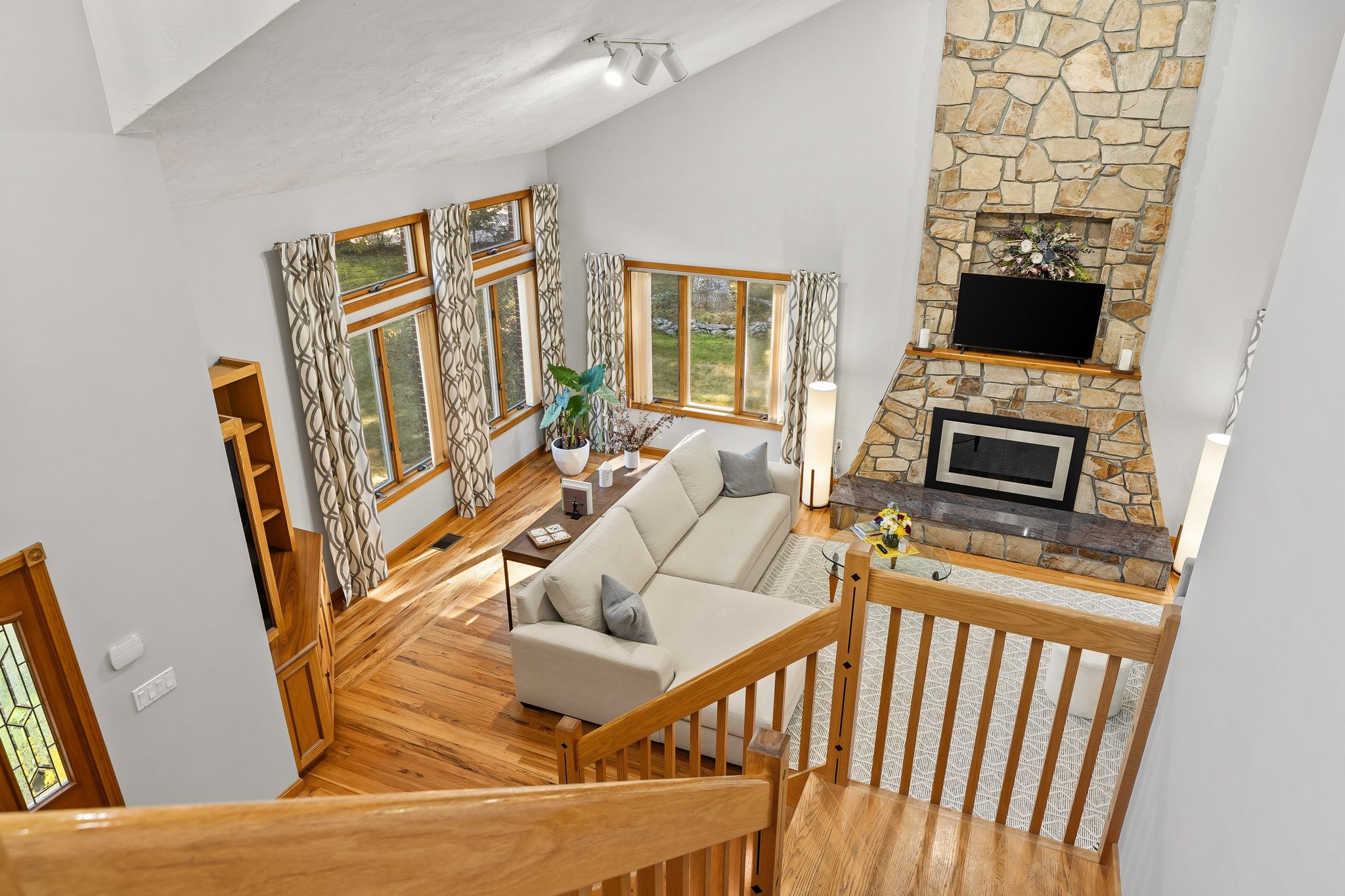 An overhead view of a bright living room featuring a stone fireplace, a sectional sofa, and large windows with hardwood floors.