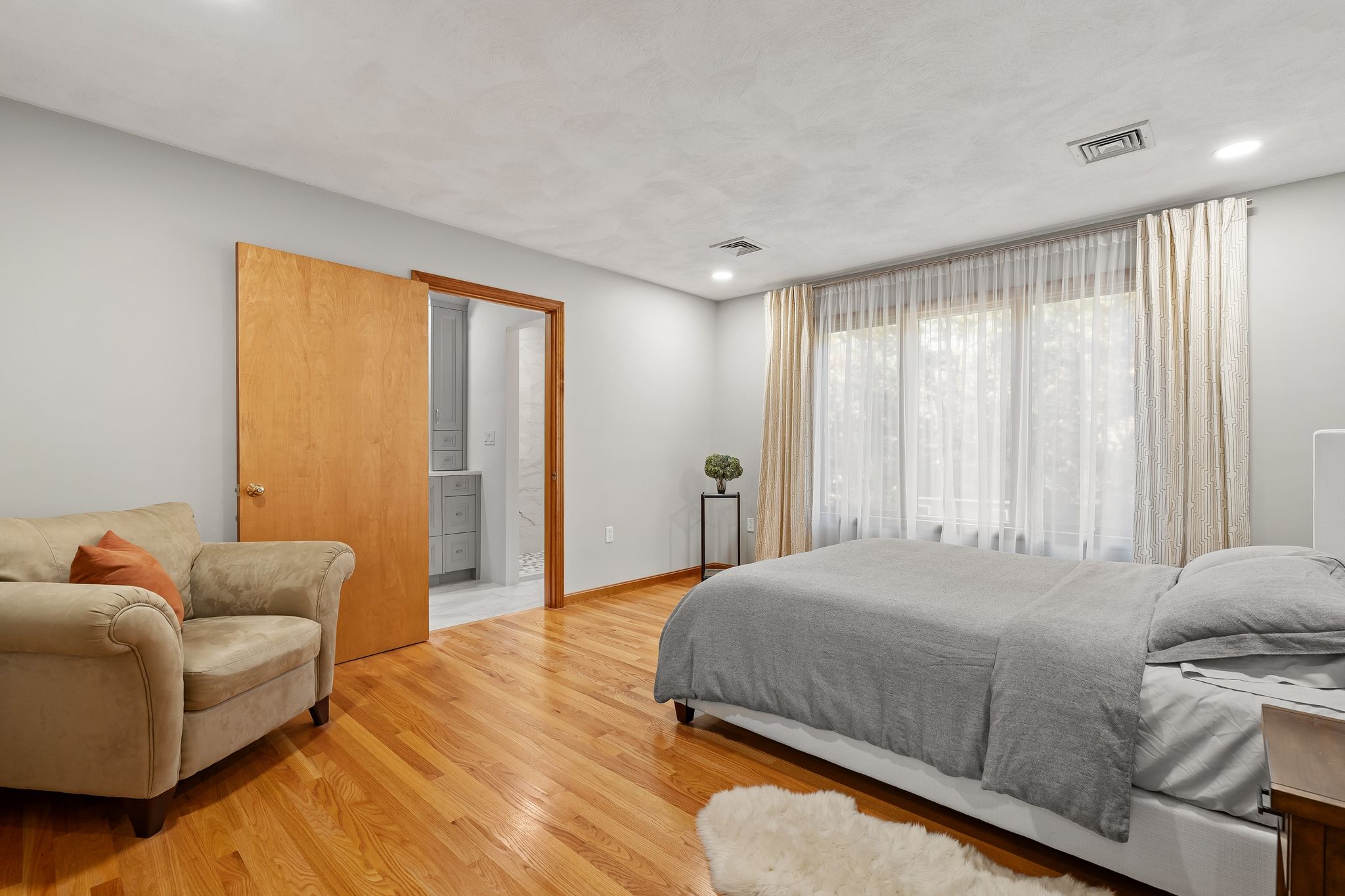 A primary bedroom with hardwood floors and large windows, featuring a bed, an armchair, and an open door leading to a bathroom.