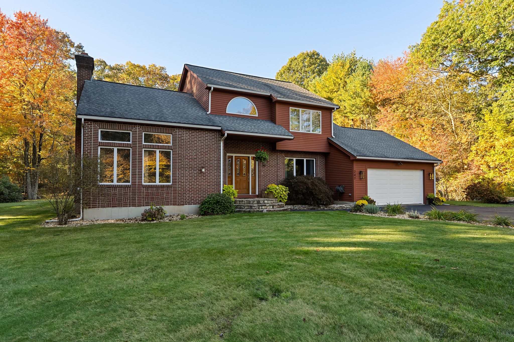 A two-story house with brick and siding exterior, a front yard, and an attached garage, set against a backdrop of autumn trees under a clear sky.