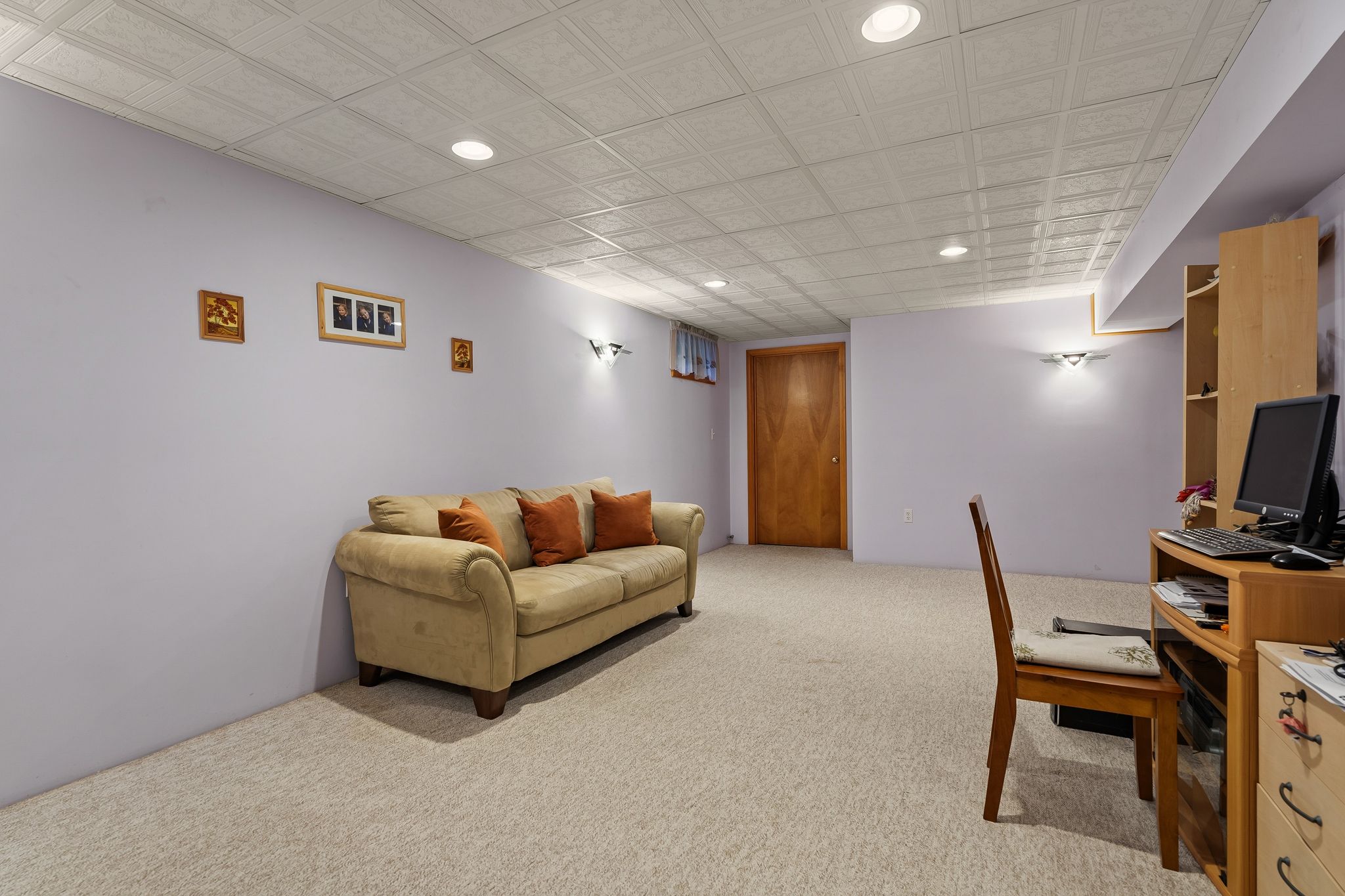 A basement living space features a light purple wall, a beige sofa, a carpeted floor, and a desk area with a computer.