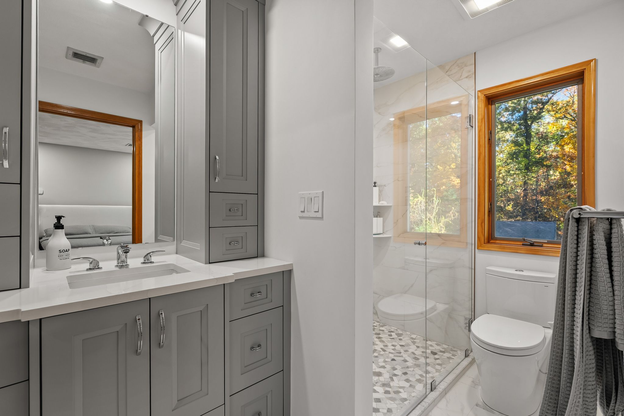 A bright bathroom features a grey vanity with a sink, a mirror, a glass shower, a toilet, and a window with a view of trees.