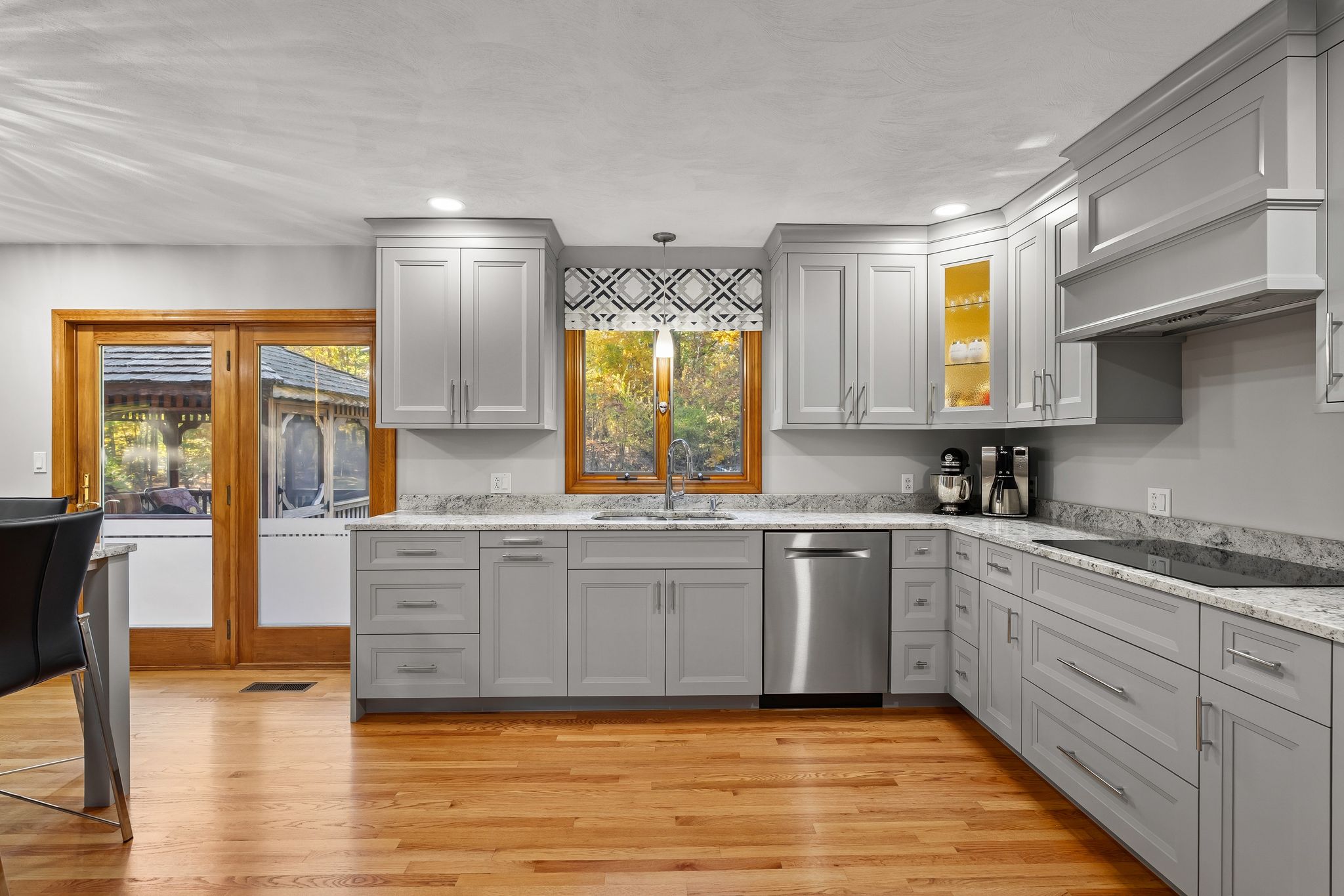 A bright kitchen features grey cabinetry, granite countertops, hardwood floors, and stainless steel appliances, with a window and glass door providing views to the outdoors.
