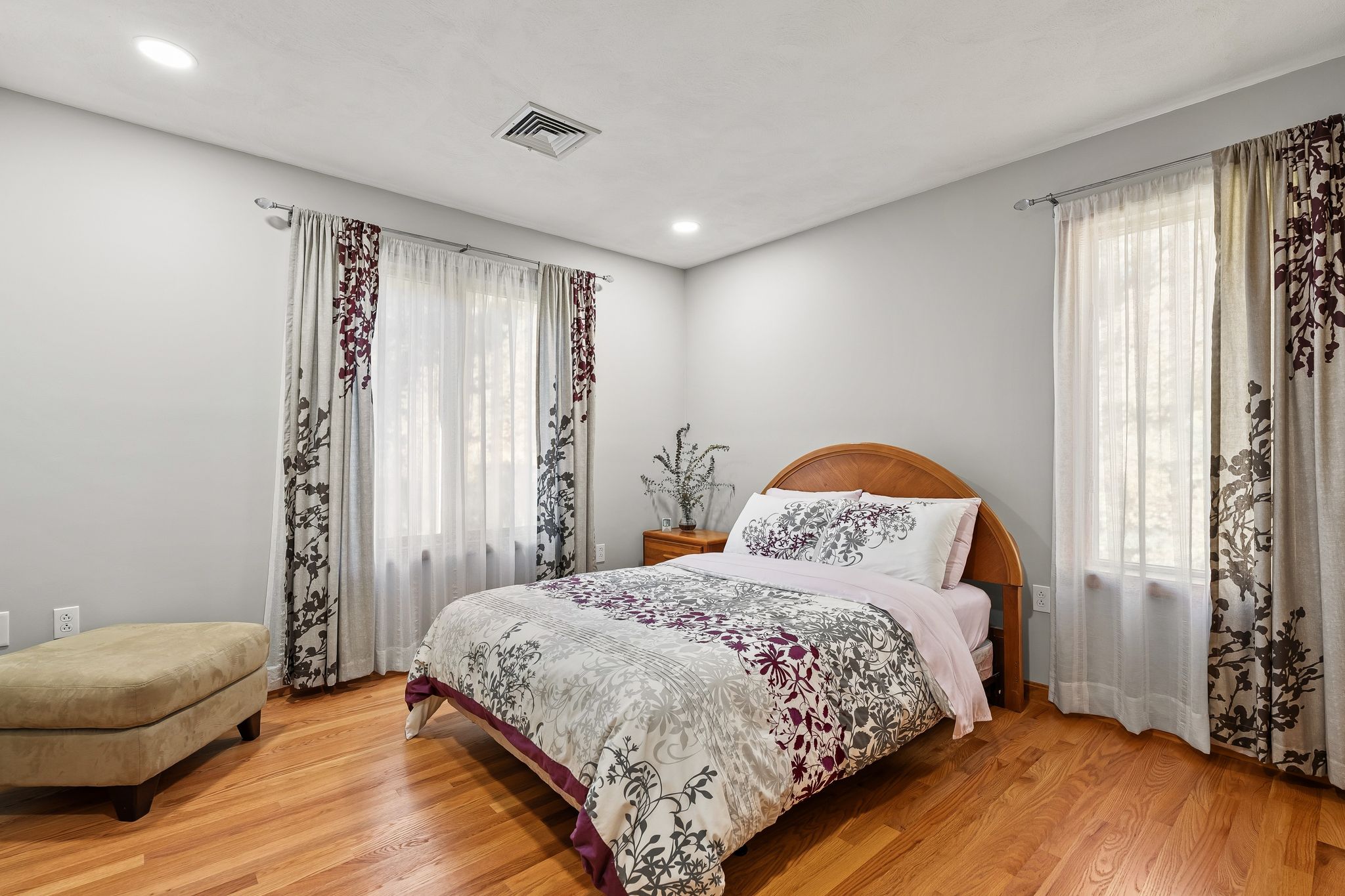 A bedroom features a bed with floral bedding, hardwood floors, and windows with patterned curtains.
