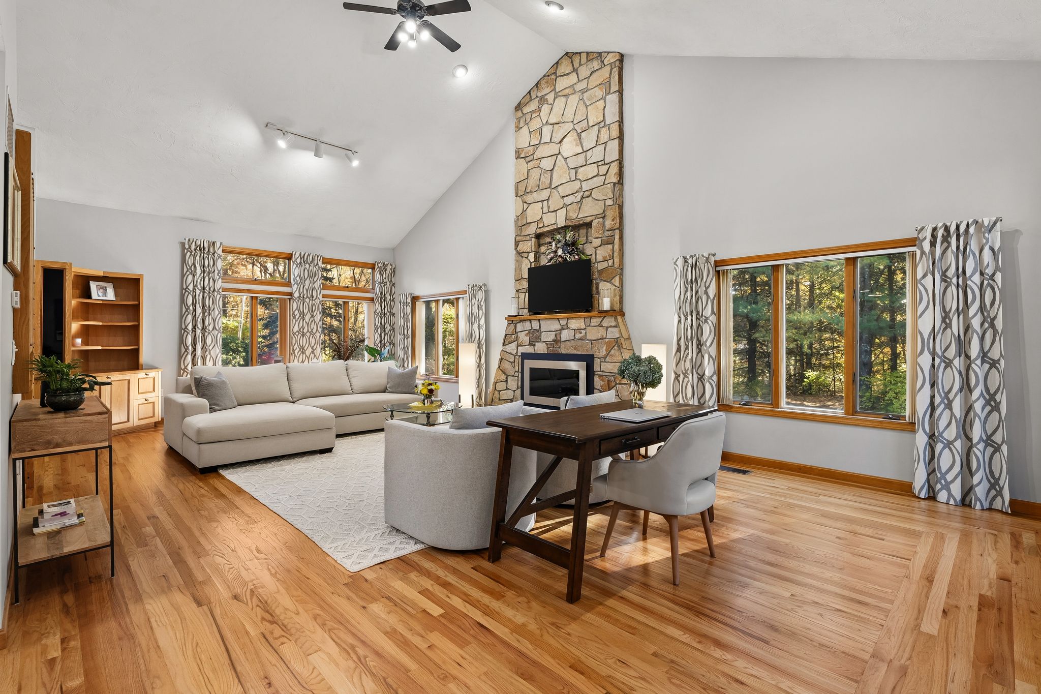 A bright living room features a stone fireplace, a large sectional sofa, and a desk area, with hardwood floors and large windows looking out to trees.