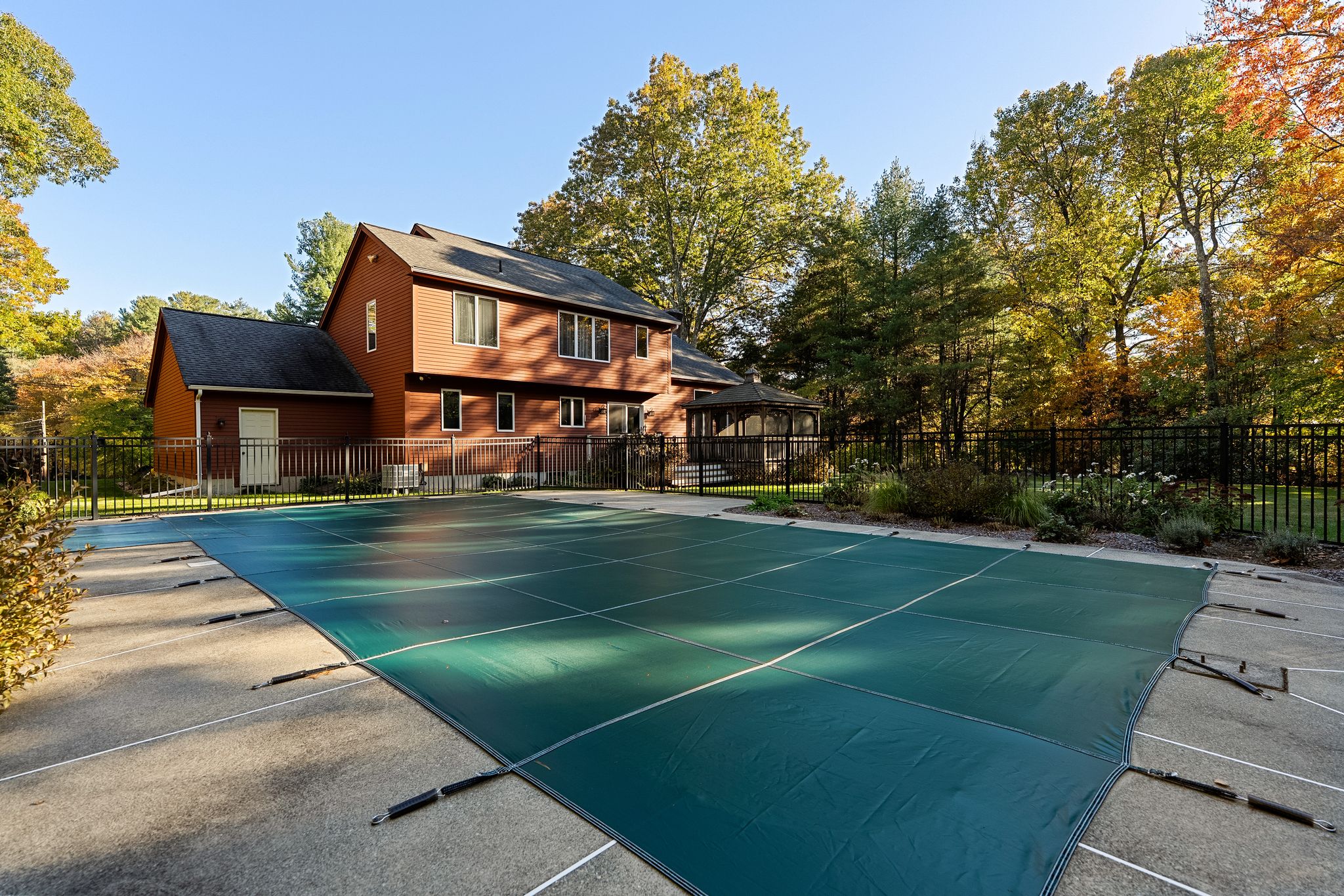 A house with a covered swimming pool and a gazebo in the backyard, surrounded by trees with autumn foliage.