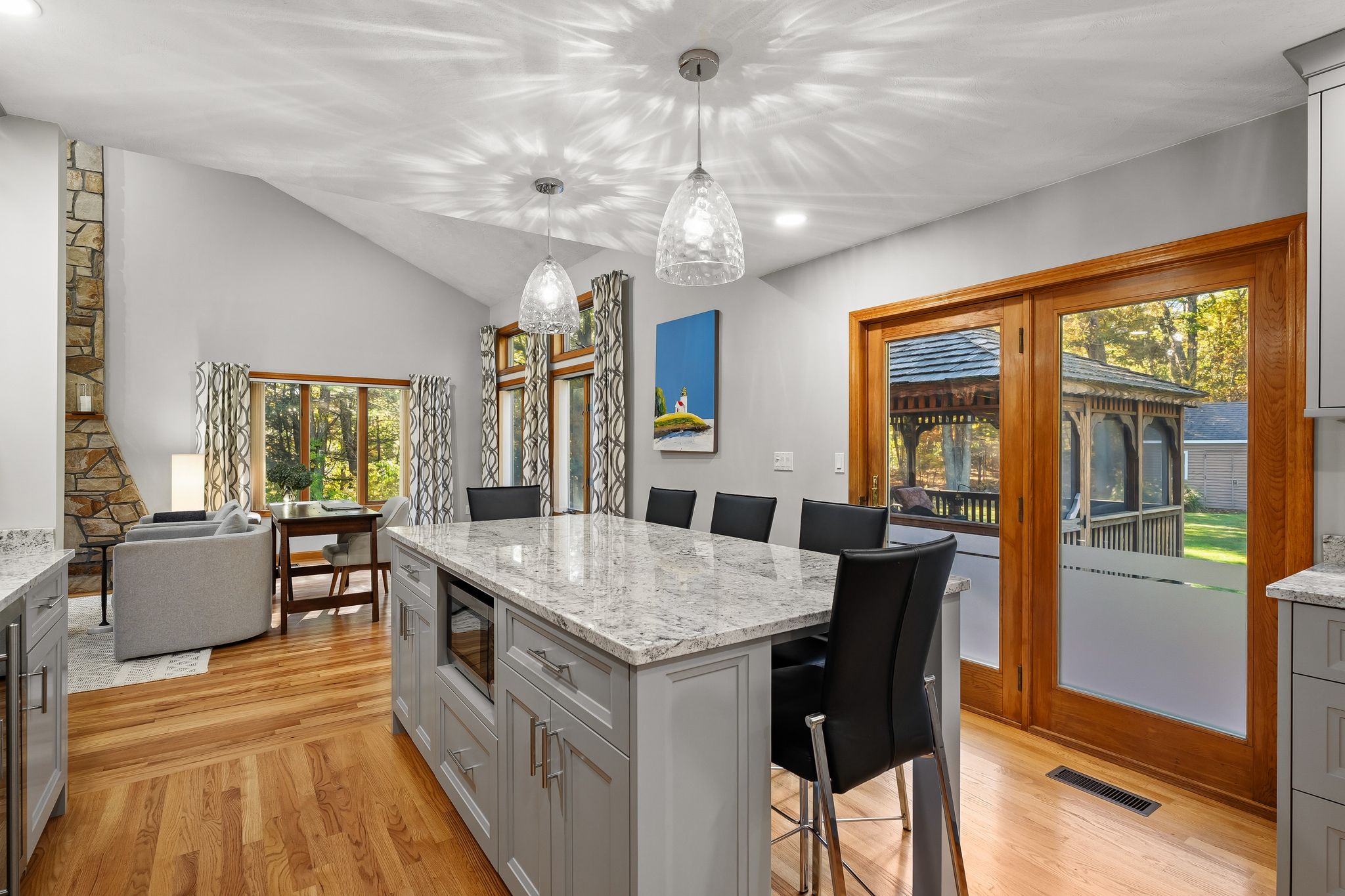 An open-concept kitchen and living room with a large island, hardwood floors, a stone fireplace, and a view to an outdoor gazebo.