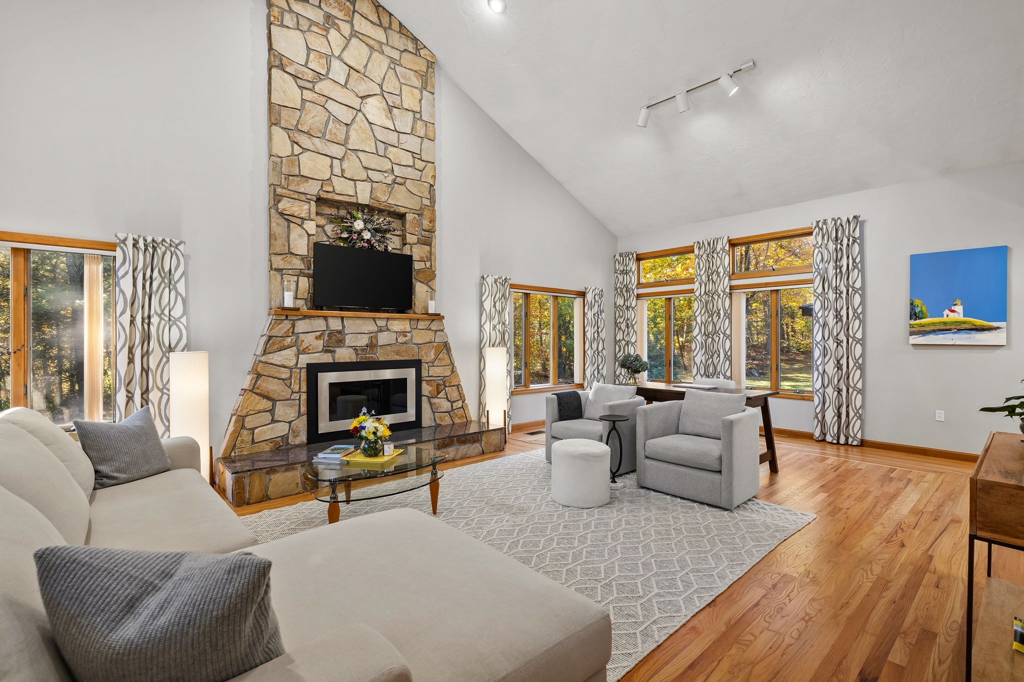 A spacious living room with a prominent stone fireplace, a large sectional sofa, armchairs, and numerous windows showcasing an outdoor view.