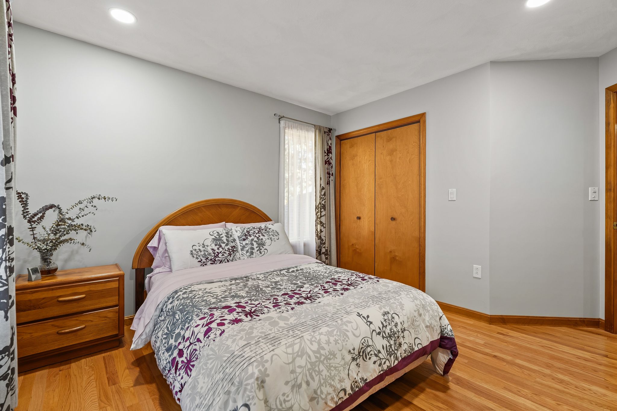 A bedroom features a bed with floral bedding, a wooden nightstand, and a wood-paneled closet with hardwood floors.