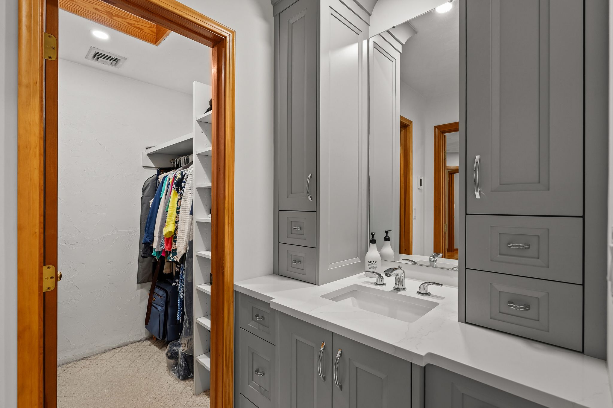 A bathroom vanity with a sink, mirror, and grey cabinets next to an open doorway leading to a walk-in closet.