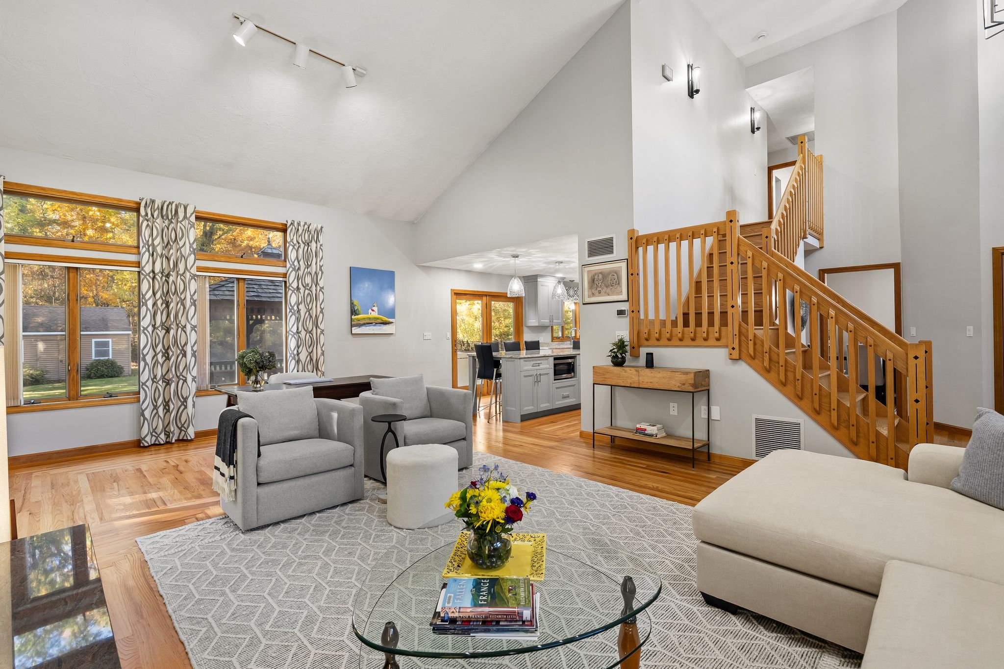 A bright living room with a vaulted ceiling, hardwood floors, a large area rug, and a wooden staircase, with a partial view of the kitchen.