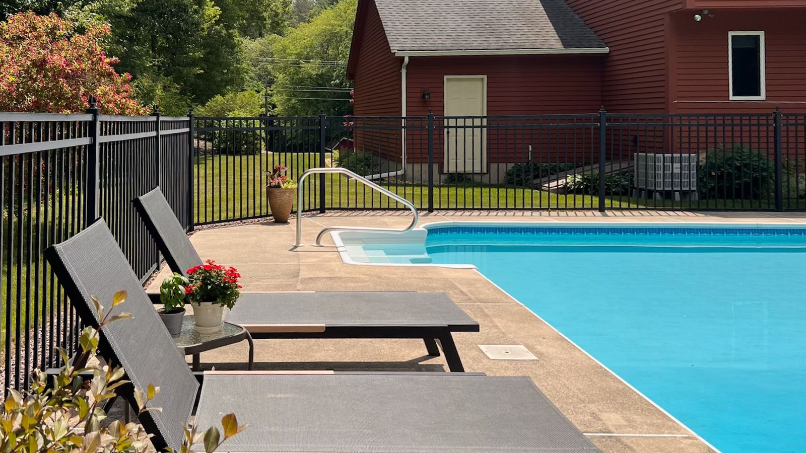 An outdoor swimming pool area with lounge chairs and a black metal fence, next to a red building.
