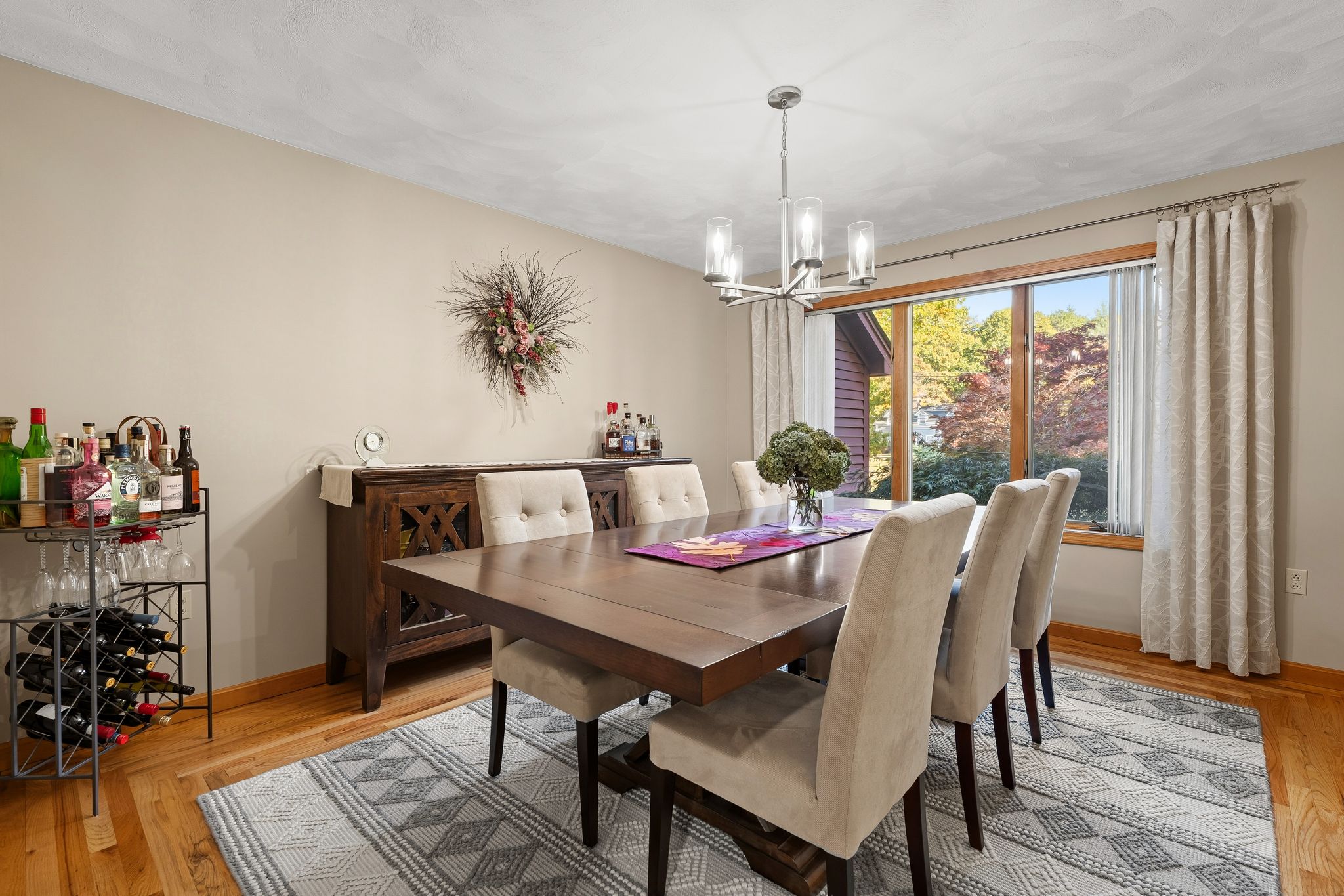 A dining room is furnished with a wooden dining table, upholstered chairs, a chandelier, a buffet cabinet, and a wine rack, next to a large window overlooking autumn foliage.