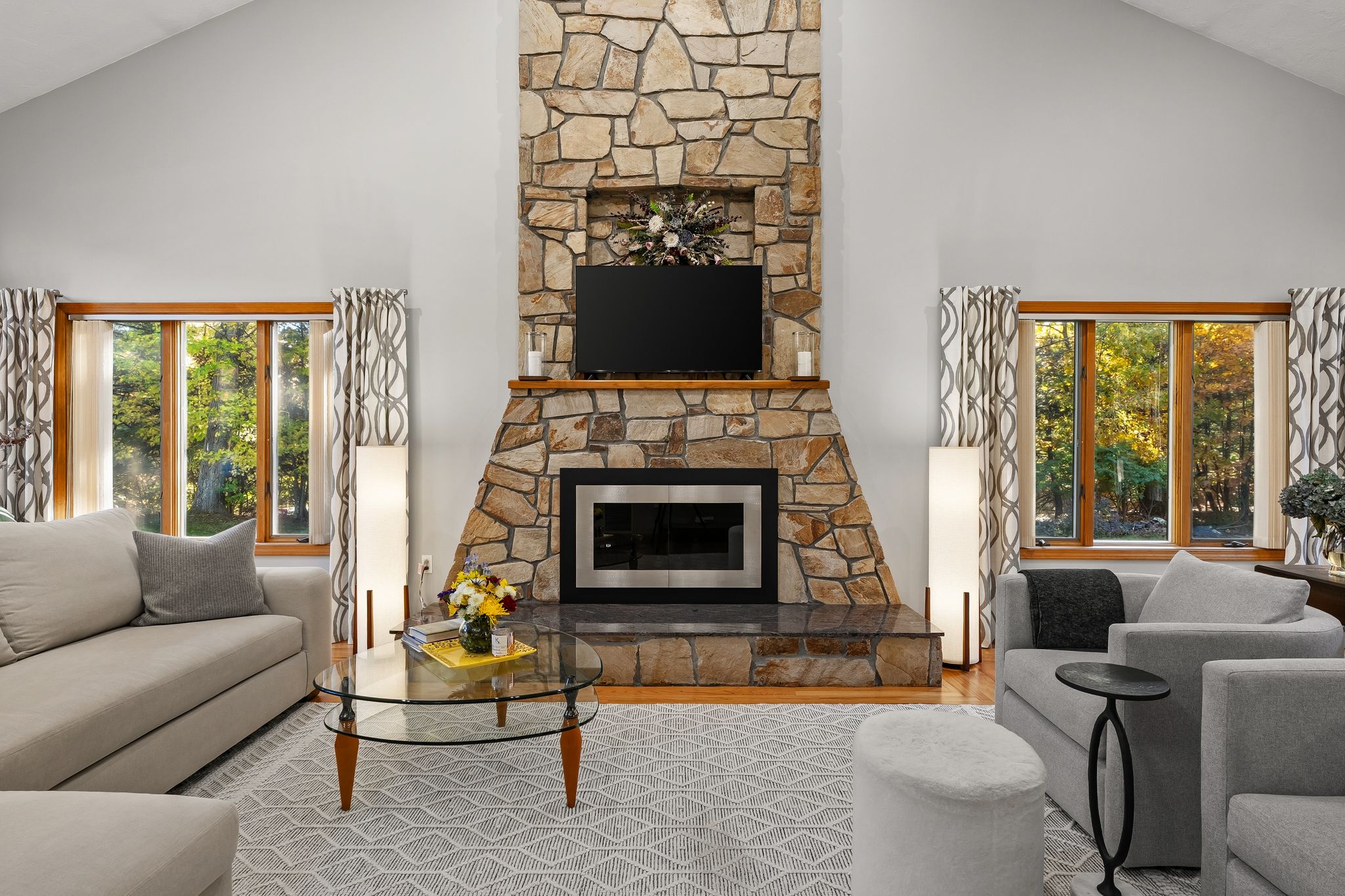 A bright living room features a prominent stone fireplace with a television above it, flanked by large windows looking out to trees.