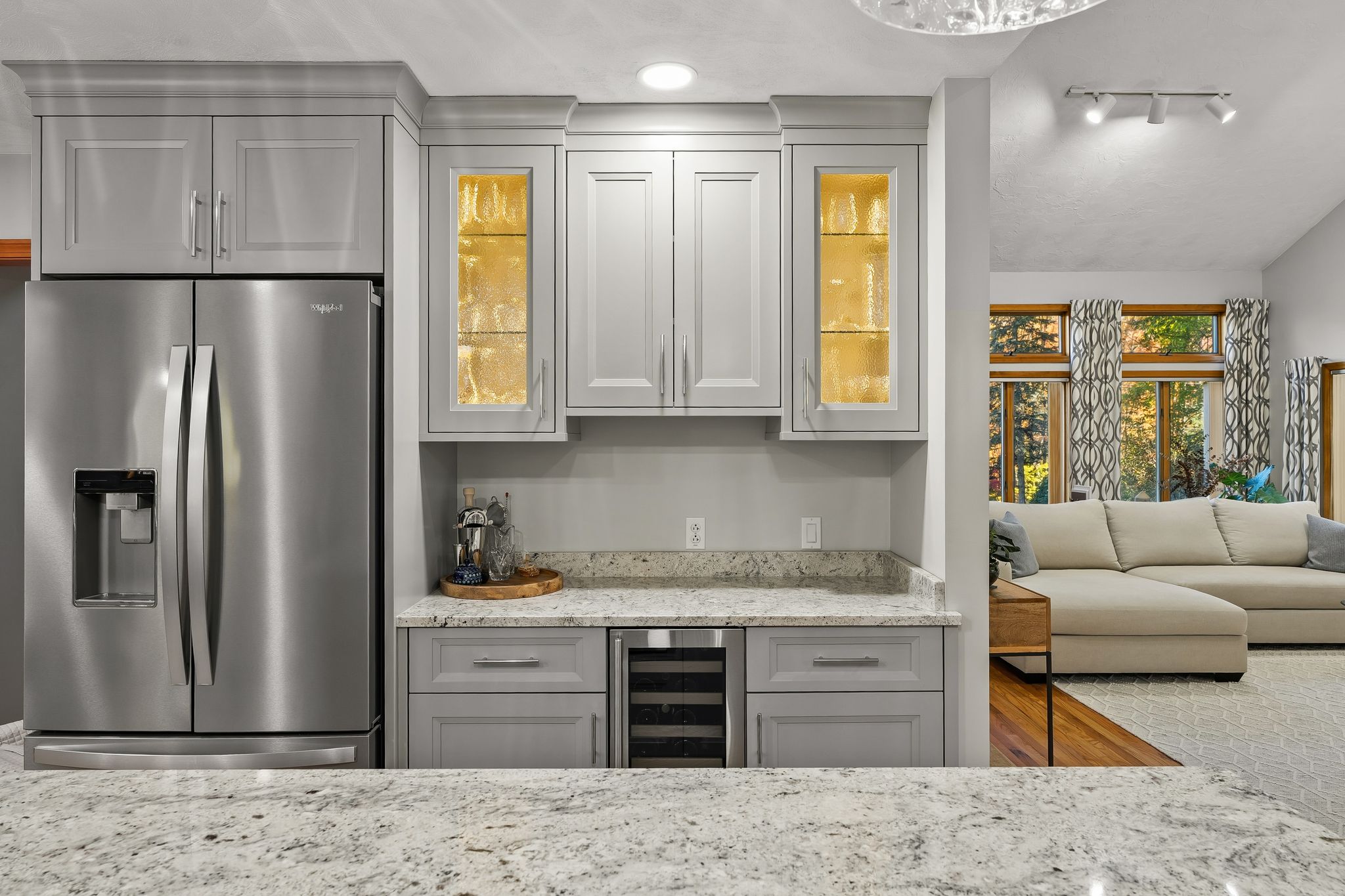 A kitchen and wet bar area with gray cabinets and granite countertops, open to a living room with large windows.