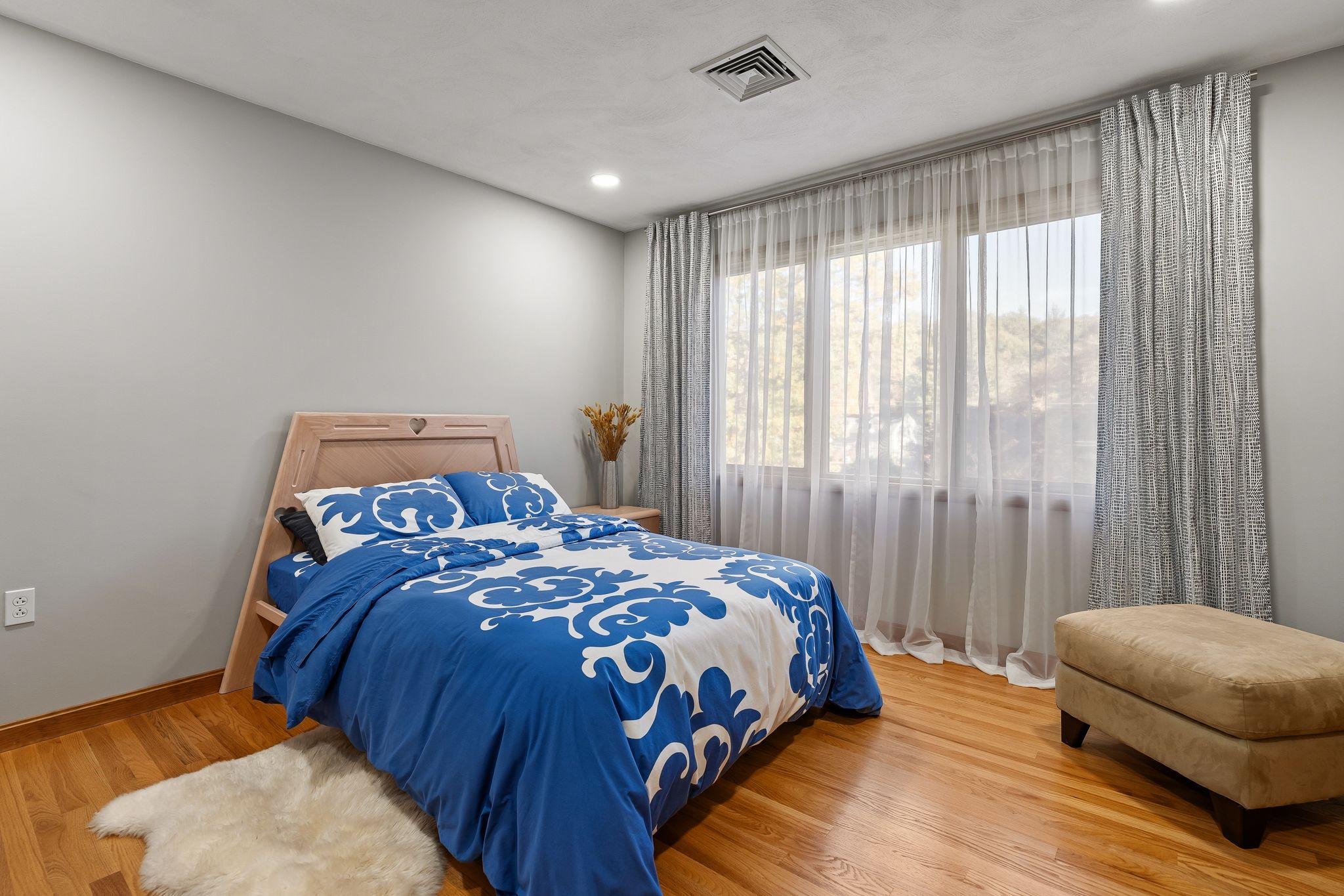 A bedroom features a bed with blue and white bedding, hardwood floors, and a large window with sheer and patterned curtains.