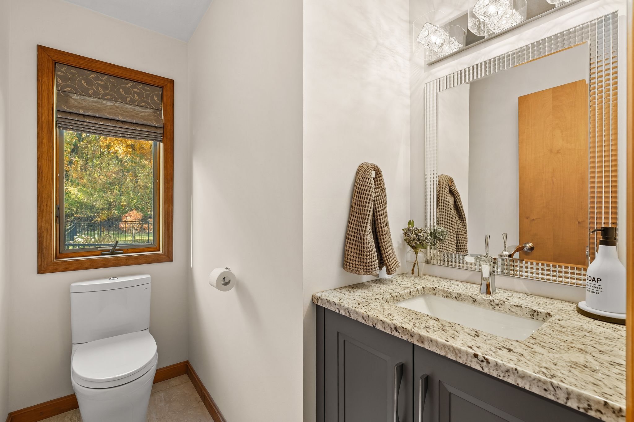 A bright bathroom featuring a toilet, a window with a view of trees, and a vanity with a sink, granite countertop, and mirror.