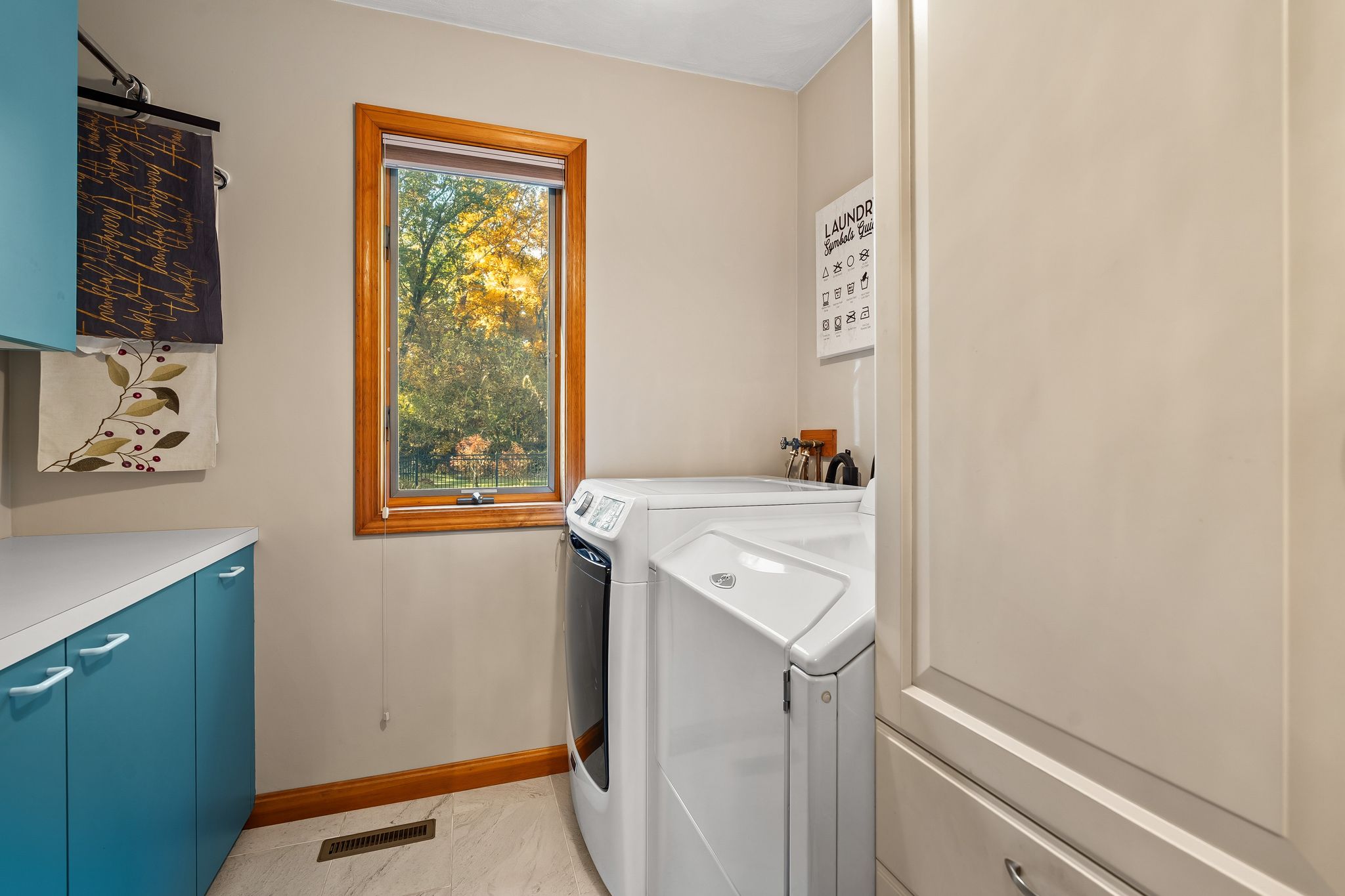 A laundry room with a washer, dryer, blue cabinets, a countertop, a window looking out to trees, and a laundry symbols guide on the wall.