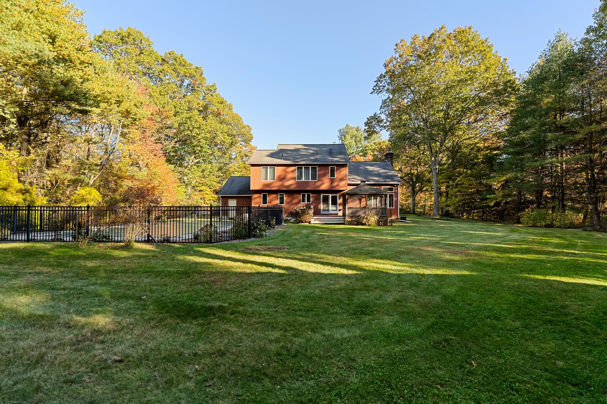 The back of a red house features a screened porch and a fenced swimming pool, set within a large green yard bordered by trees with autumn foliage.