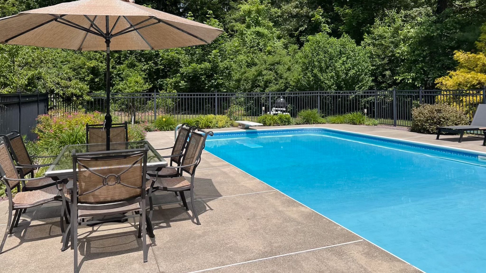 An outdoor swimming pool with a patio featuring a dining set and umbrella, surrounded by a fence and trees.