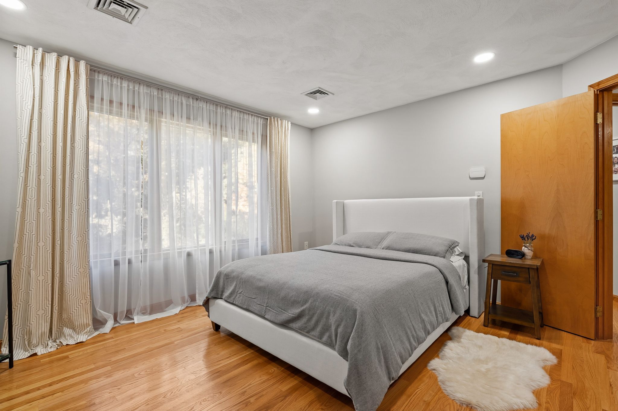 A bright primary bedroom features hardwood floors, a white upholstered bed with grey bedding, large windows with sheer and patterned curtains, and a wooden nightstand.