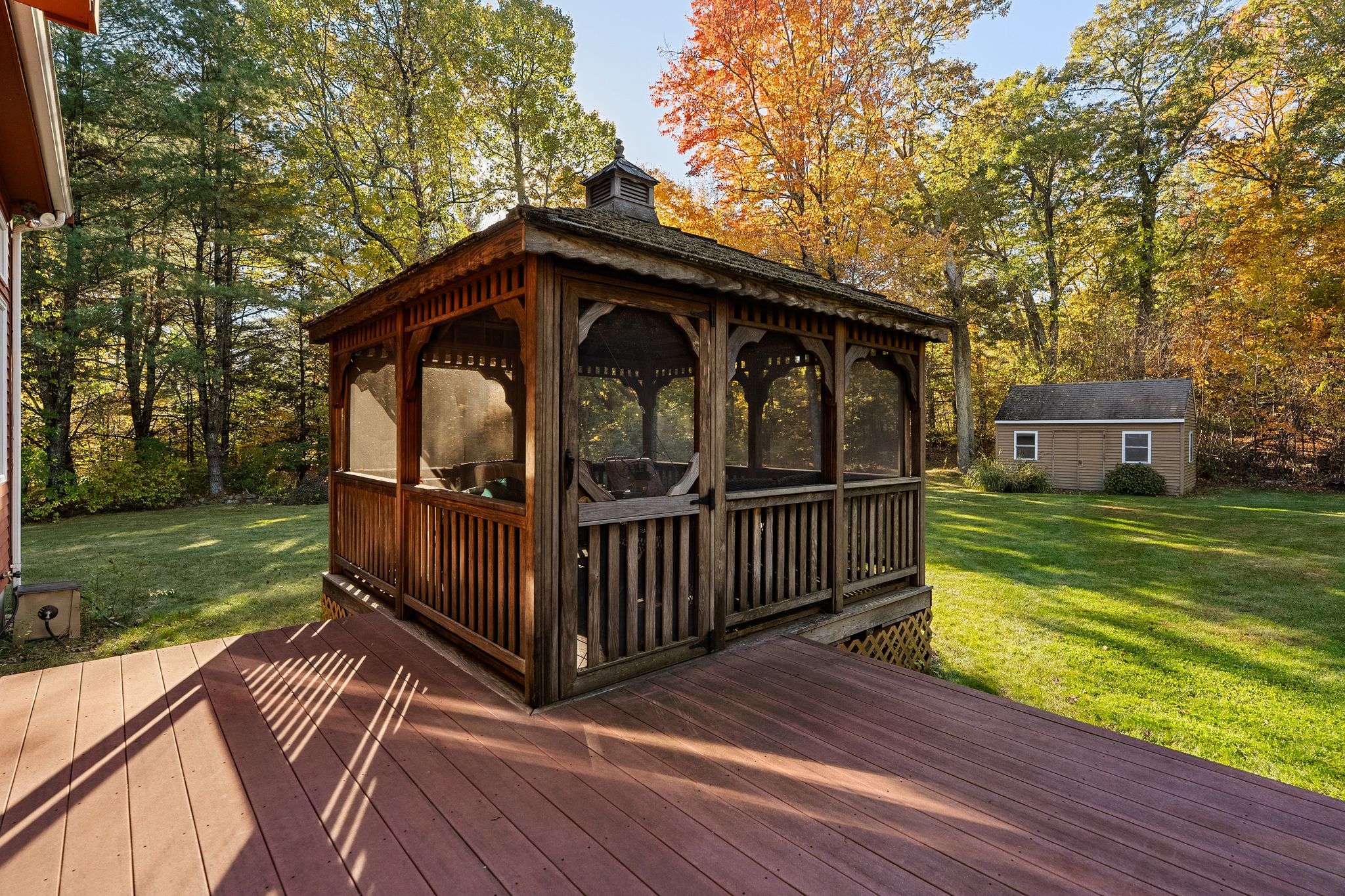 A wooden gazebo is on a deck in a sunny backyard with autumn trees and a shed in the background.