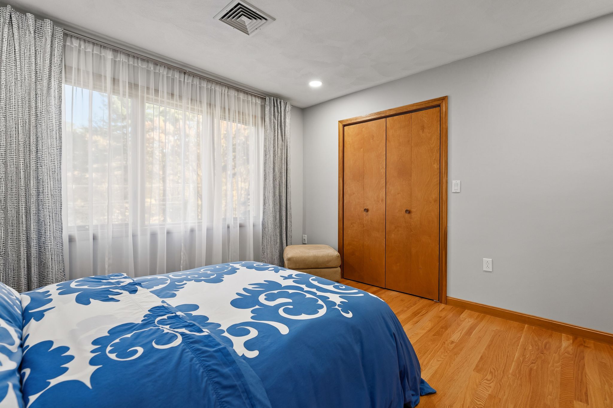 A bedroom features a bed with blue and white bedding, hardwood floors, a large window with sheer curtains, and a closet.